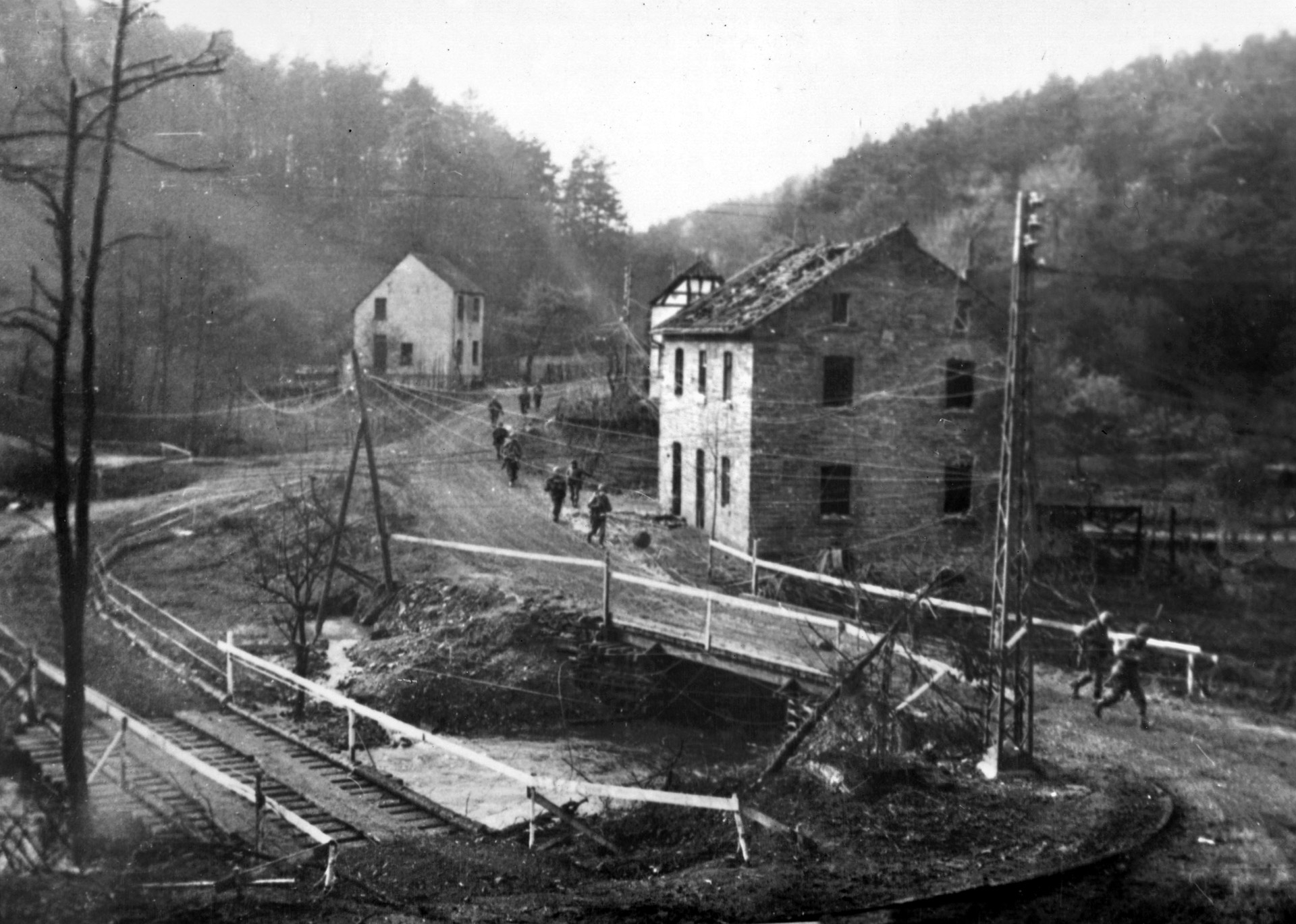 A line of American troops crosses a bridge and marches through one of the many small villages that dot the Hürtgen region. The difficult hilly and forested terrain is very obvious in this photo, taken November 16, 1944.